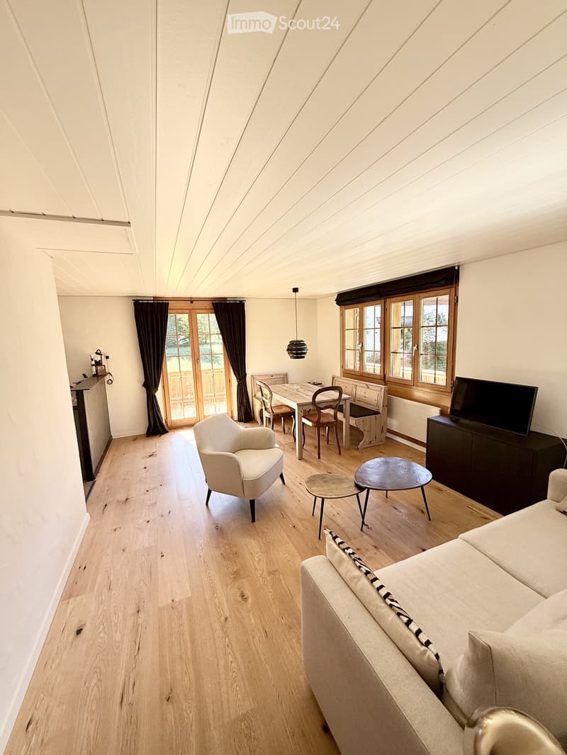 Bright living room with wooden flooring, white walls, and a slanted wooden ceiling. Features a cream-colored sofa, a white armchair, a coffee table, a dining area with a table and chairs, and a television on a stand.