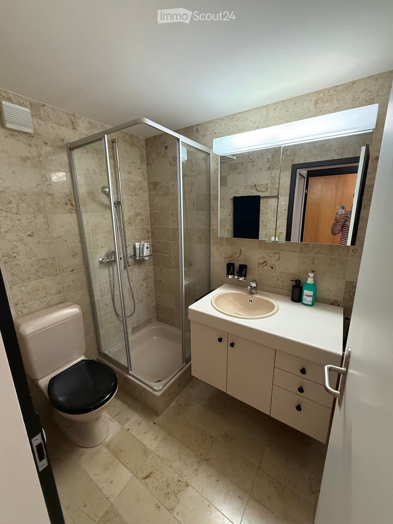 Modern bathroom with beige tiles, a glass-enclosed shower, a toilet, a sink with a mirror, and a cabinet.
