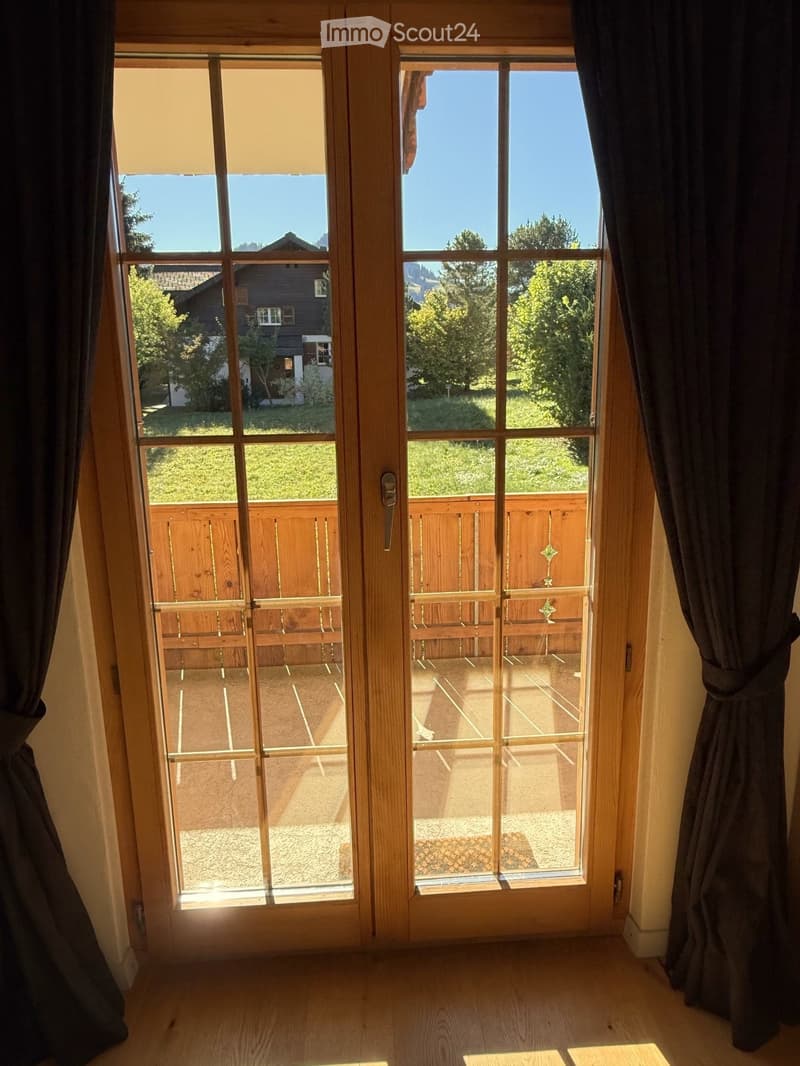 View through large wooden-framed glass doors showing a wooden deck with a railing and a view of a green landscape and neighboring house.