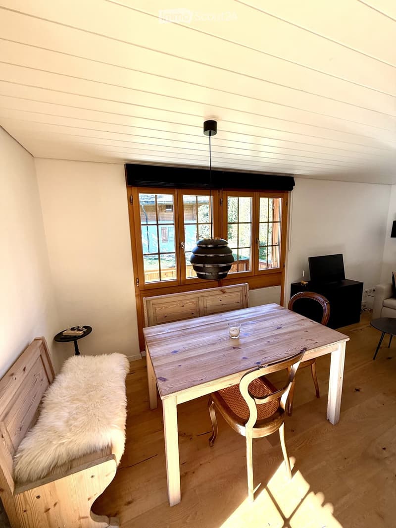 Brightly lit room with wooden floor, large window, wooden bench with fur throw, dining table with chairs, small black stool, and TV on stand.