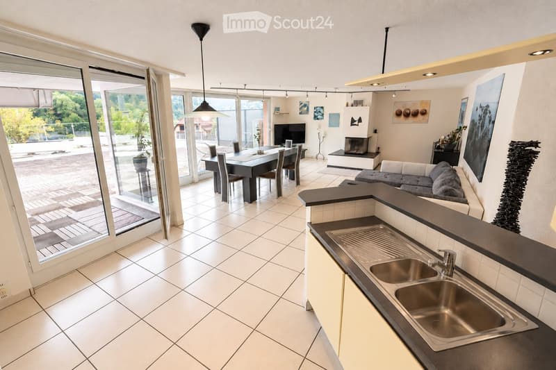 Open-plan living and dining area with modern furniture, large windows, and a kitchen island with a sink.