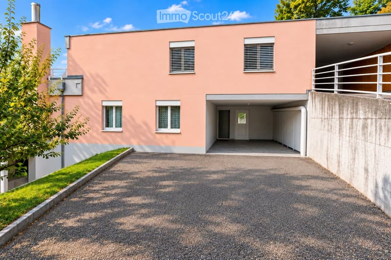 Modern two-story house with pink facade, white-framed windows, and a covered garage entrance. The driveway is paved, and there is a small garden area with green grass and a tree on the left side.