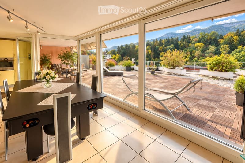 Bright dining area with a modern black dining table, white chairs, and a vase of white flowers. Large glass doors open to a wooden deck with lounge chairs and a scenic mountain view.