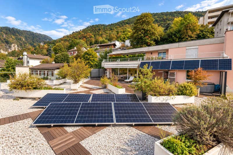 A modern rooftop garden with solar panels, wooden decking, potted plants, and lounge chairs, overlooking a scenic hillside with trees and houses.