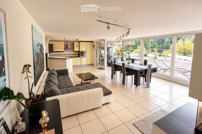 Modern living and dining area with a grey sectional sofa, glass coffee table, dining table with chairs, and a small kitchen area with cabinets and appliances.
