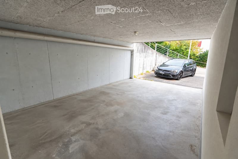 Underground garage with concrete walls and ceiling, one parked car visible, natural light from an opening at the far end.