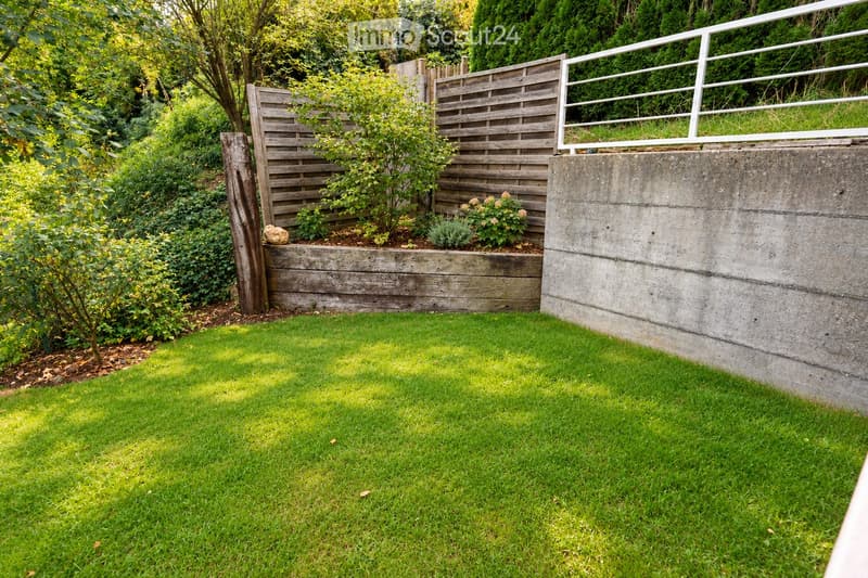 Well-maintained garden with a concrete retaining wall, wooden fence, small plants, and lush green grass.