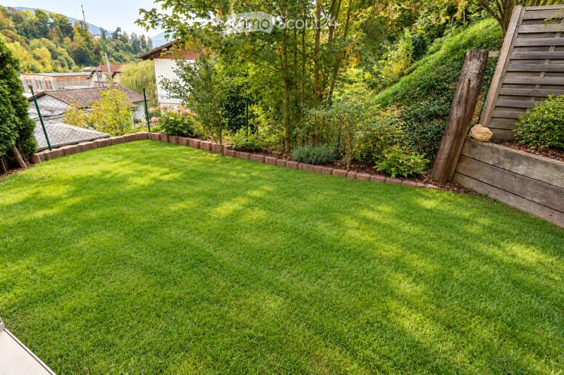 Well-maintained lawn with neatly trimmed grass, surrounded by a low stone border, small plants, and trees. Wooden fence and wooden post visible on the right side.
