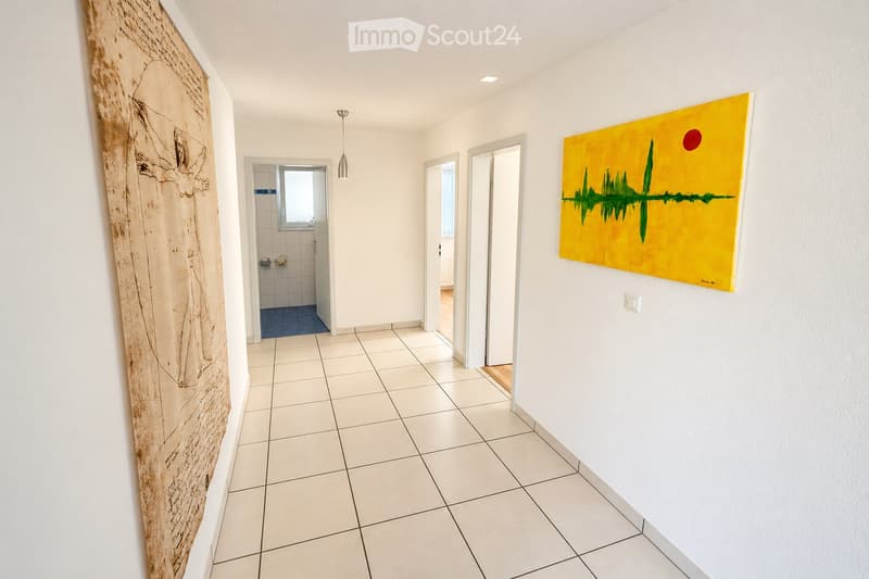 Bright hallway with white tiled flooring, white walls, and modern lighting. Features a large abstract painting on one wall and a doorway leading to a tiled bathroom with blue accents.