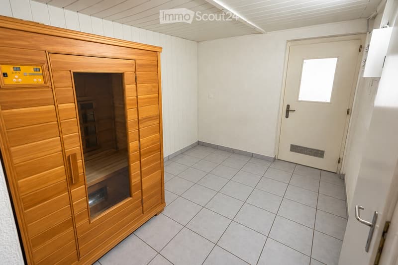 A small, tiled room with a wooden sauna unit featuring a control panel and glass door. The room has a white door with a small window and a tiled floor.