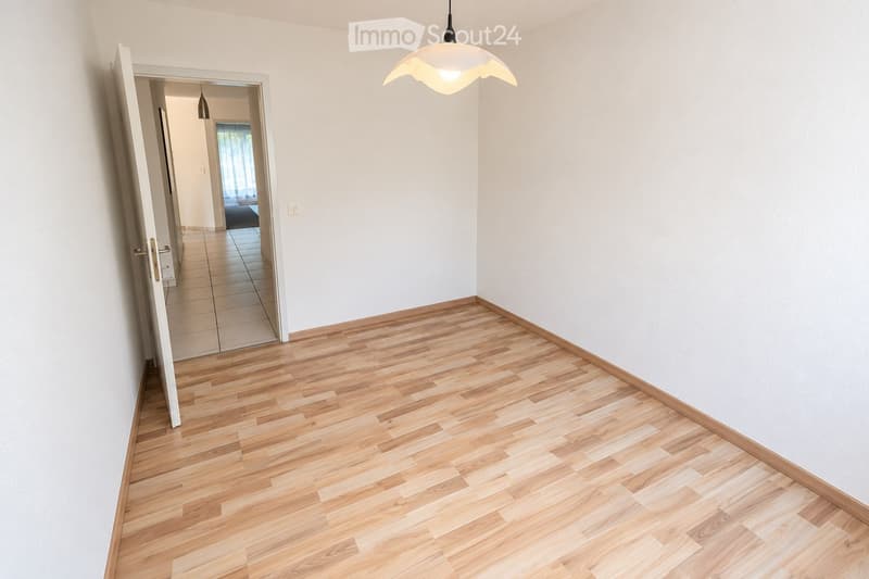 Empty hallway with light wooden flooring, white walls, and a hanging light fixture. An open door leads to another room with tiled flooring.