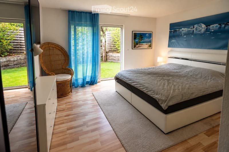 Modern bedroom with a large bed, wicker chair, nightstand, and artwork on the walls. The room features wooden flooring and large windows with blue curtains, offering a view of a green garden.