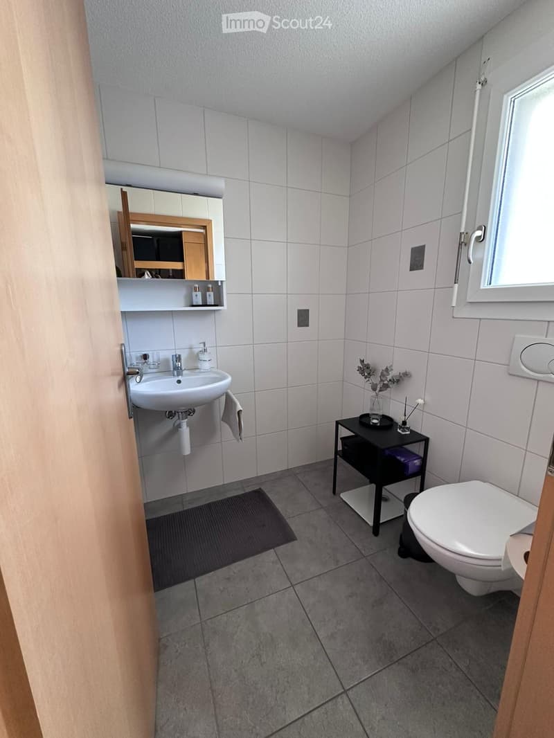 Modern bathroom with white tiles, a sink, mirror, toilet, small black stool, and a decorative vase on the stool.