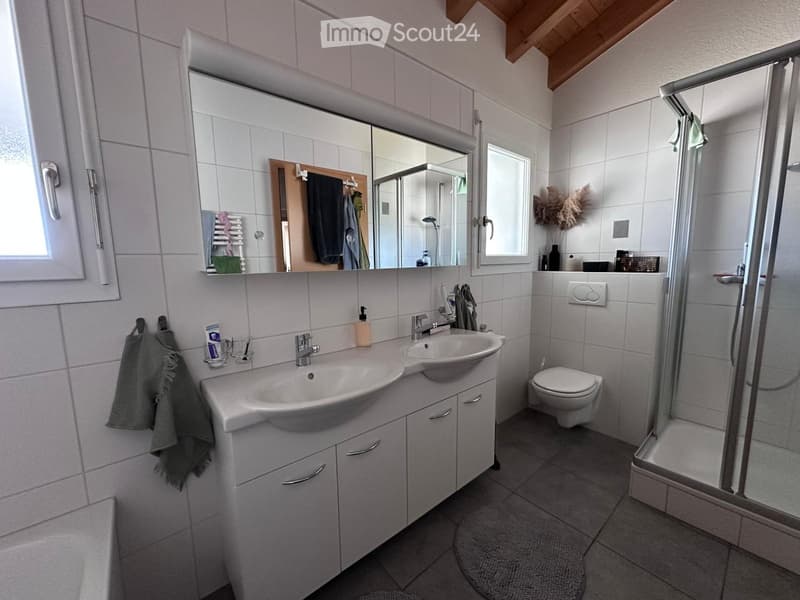 Modern bathroom with two sinks, a toilet, a shower, and a large mirror above the sinks. The room features white tiles, a wooden ceiling, and a small window.