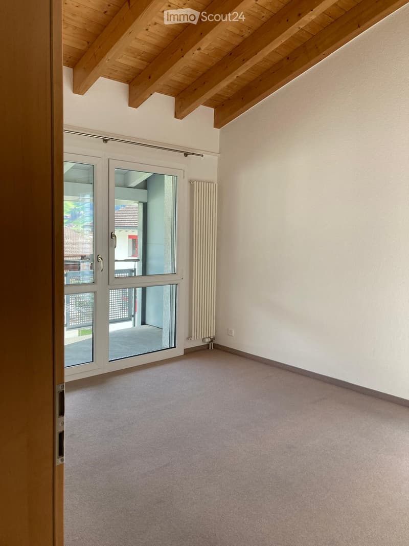 Empty room with light-colored walls, carpeted floor, large windows with white frames, and a radiator on one wall.