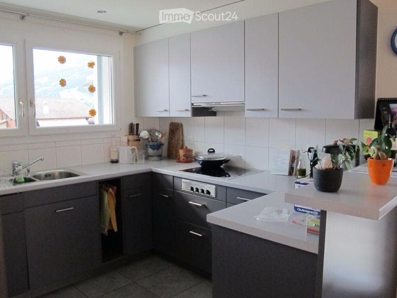 Modern kitchen with light grey cabinets, white countertops, and stainless steel appliances. Features a sink, stove, oven, and various kitchen items on the counter.