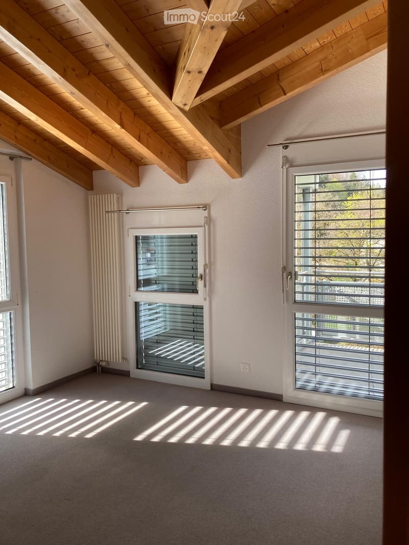 Empty room with exposed wooden beams on the ceiling, large windows with blinds, and a radiator on the wall.