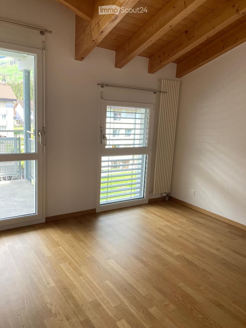 Empty room with wooden flooring, white walls, large windows, and a sliding door leading to a balcony.