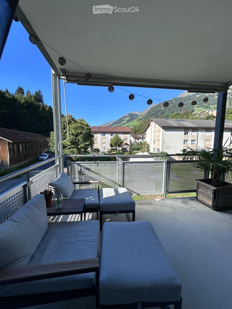 Balcony with a view of mountains, featuring a sofa, two stools, a small table with a cup, and potted plants.