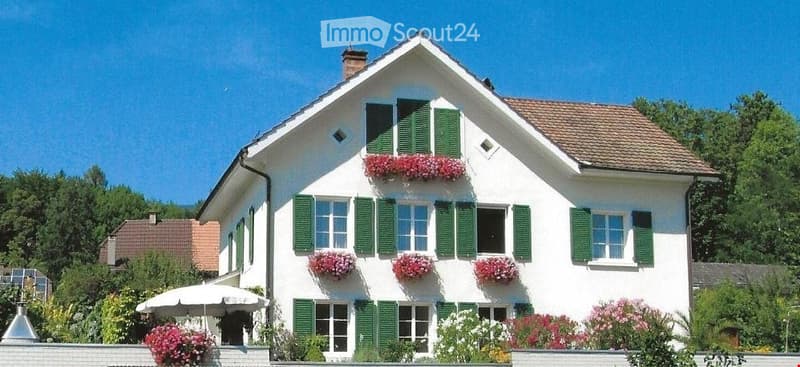 Two-story white house with green shutters, multiple windows, and flower boxes on the facade. The house has a brown tiled roof and is surrounded by greenery and a small garden area with an umbrella.