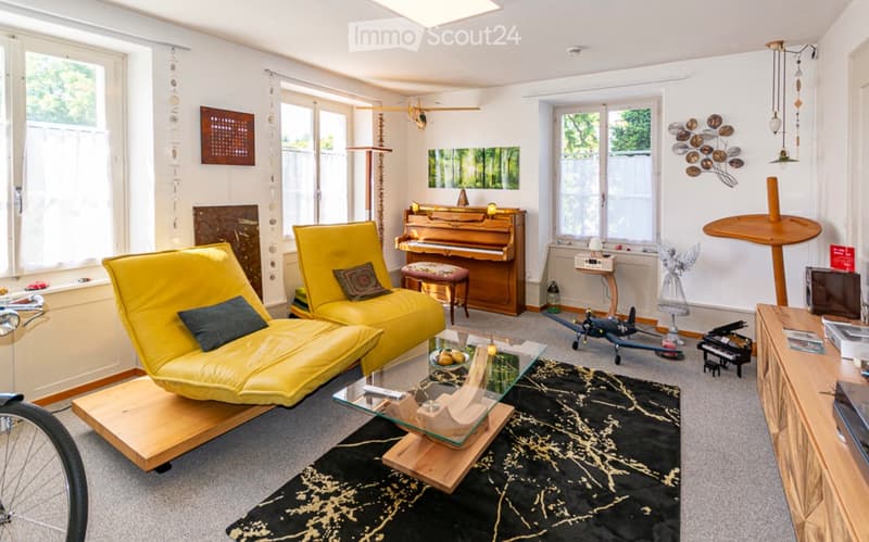 Bright living room with large windows, yellow sofa, glass coffee table, piano, and various decorative items including a wall-mounted art piece and a hanging light fixture.