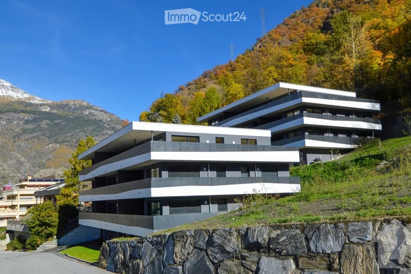 Modern multi-story building with clean lines and large windows, situated on a hillside with a scenic mountain backdrop.