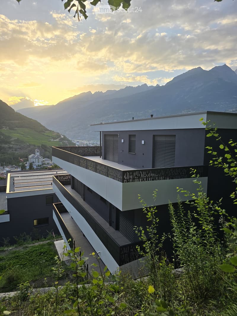 Modern two-story house with grey and white exterior, large windows, and a balcony overlooking a scenic mountain valley.