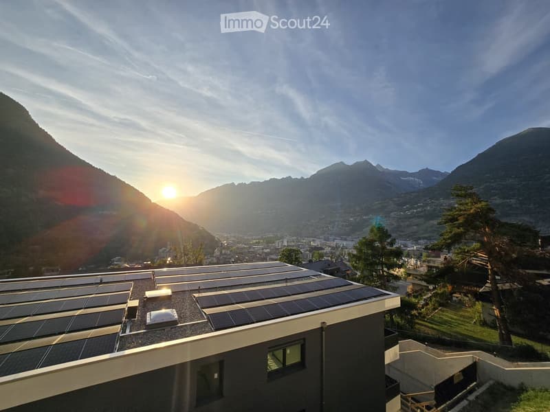 Modern building with solar panels on the roof, overlooking a scenic mountain valley and town during sunset.