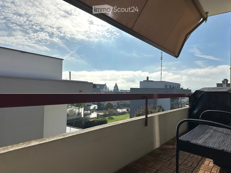 Balcony with wooden decking, metal railing, and a covered awning, featuring a single chair and a small table.