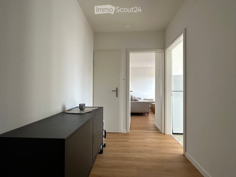 A narrow hallway with wooden flooring, white walls, and a black console table with a cup on it. The hallway leads to an open living area with a sofa and a coffee table visible in the background.