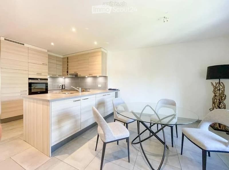Modern kitchen with light wood cabinetry, a central island, and a glass dining table with four chairs. The space includes an oven, sink, and a floor lamp near the dining area.