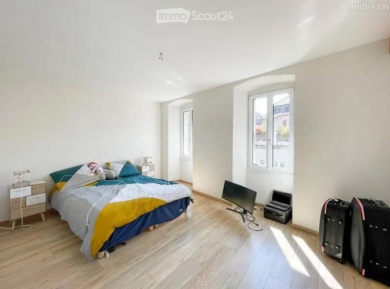 Bright bedroom with wooden flooring, large windows, a neatly made bed with colorful bedding, a nightstand, a laptop, a screen, and two suitcases near the window.
