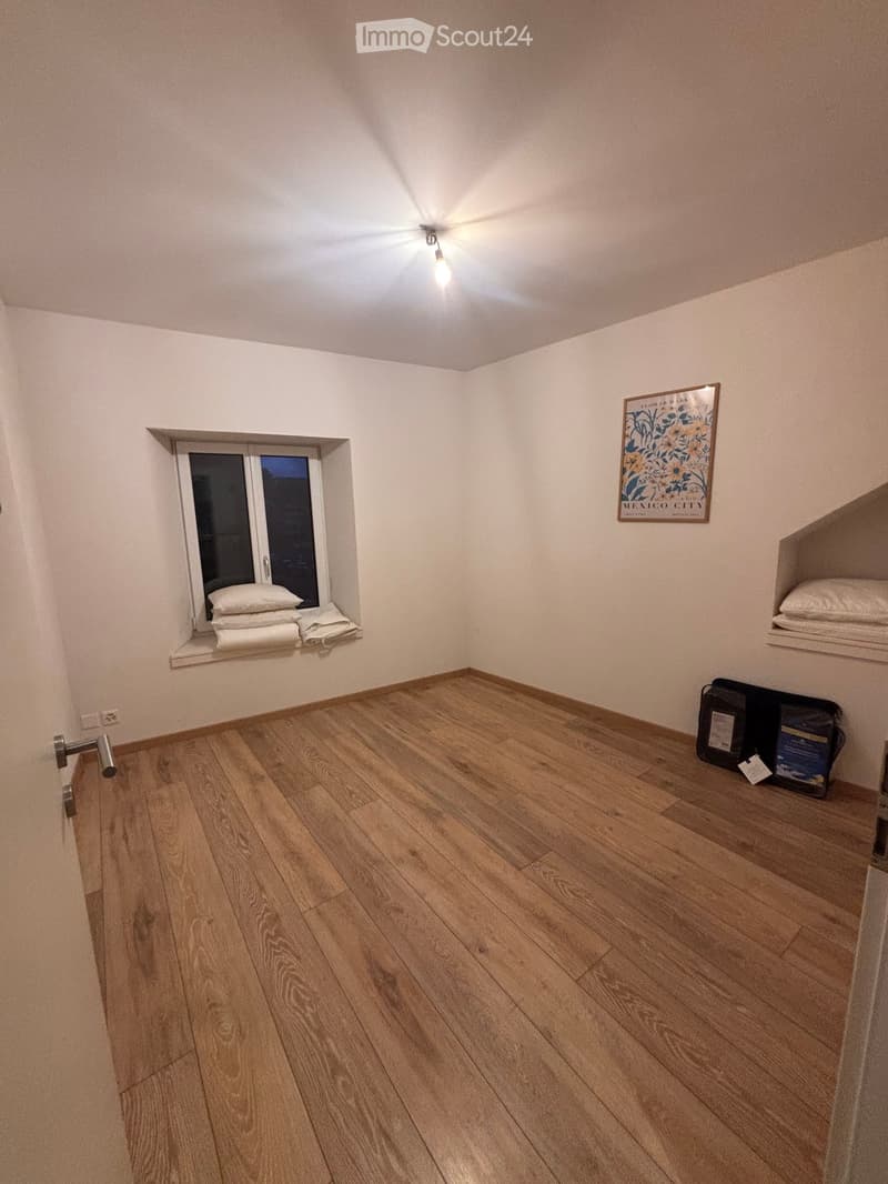 Empty room with wooden flooring, white walls, a small window with folded towels, a built-in shelf with folded towels, and a few bags on the floor.