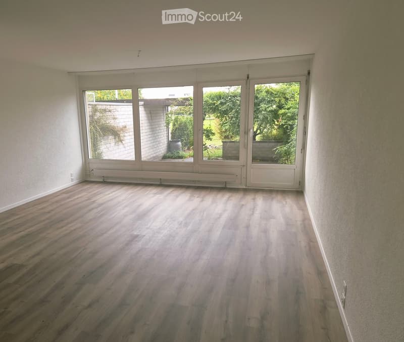 Empty room with wooden flooring, large sliding glass doors leading to a garden, and white walls.