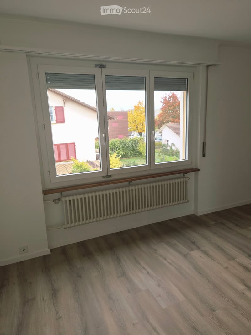 Empty room with wooden flooring, large window with blinds, radiator, and view of neighboring houses and trees.