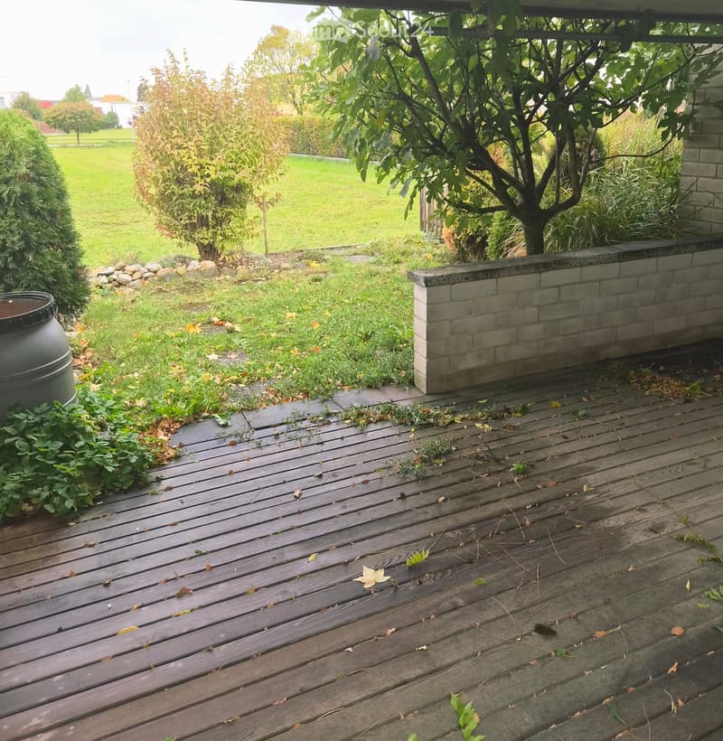 Wooden deck with scattered leaves, surrounded by a low brick wall, grassy area, shrubs, and a tree.