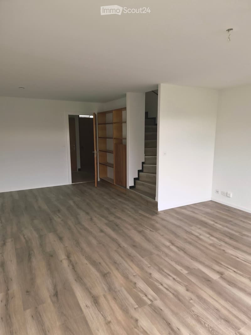 Empty room with wooden flooring, white walls, and a staircase leading upwards. There is a partially open door leading to another room and a built-in wooden shelf unit.