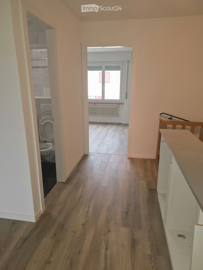 Empty hallway with wooden flooring, light-colored walls, and a view into a bathroom and another room with a window and radiator.