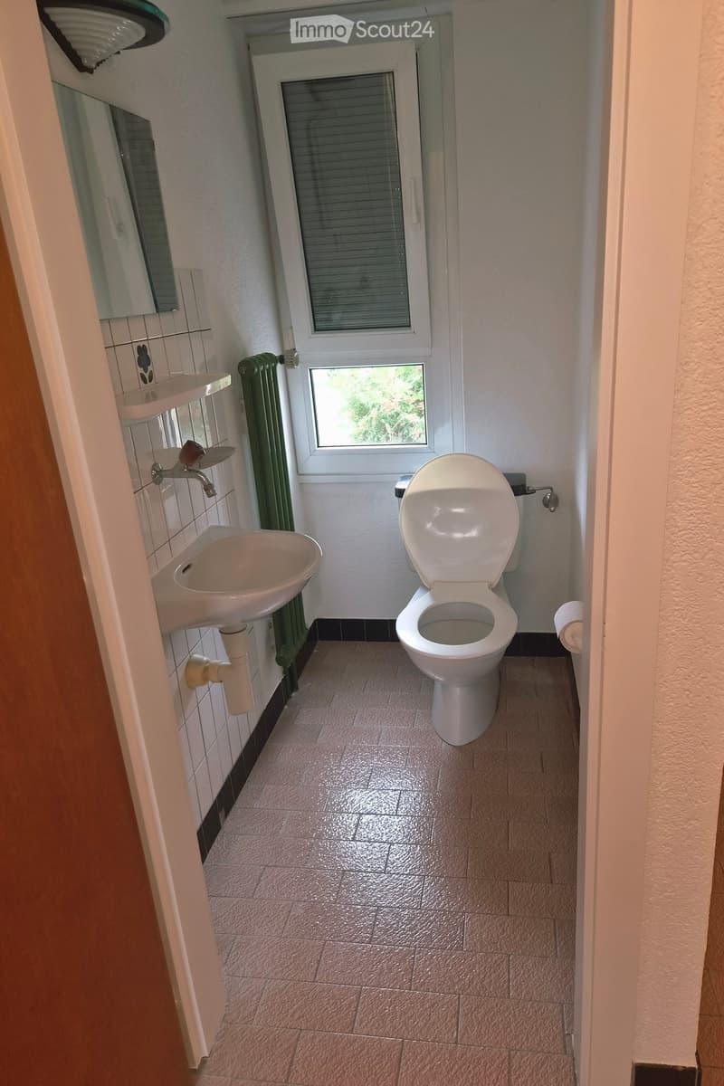 A small bathroom with a toilet, sink, radiator, and a window with blinds. The walls are tiled, and the floor is covered with light-colored tiles.