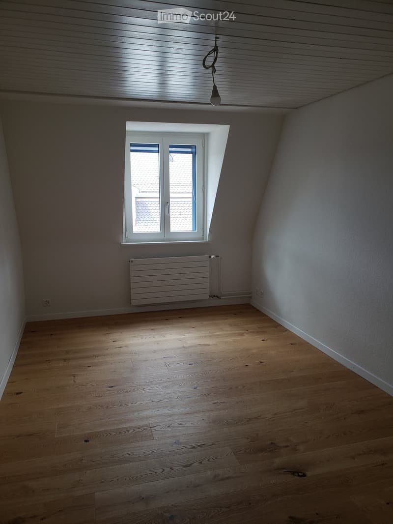 Empty attic room with wooden flooring, white walls, a single window, and a radiator. The ceiling has exposed wooden beams and a hanging light fixture.