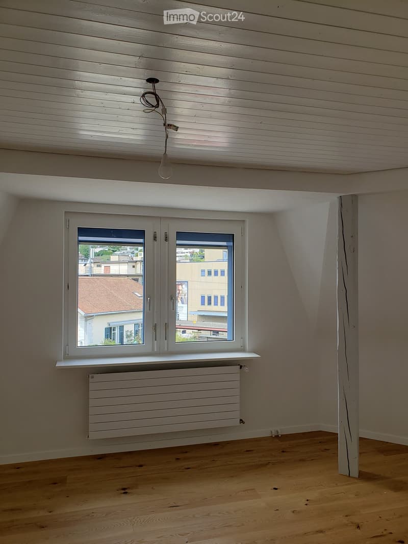 Empty room with wooden flooring, white paneled walls, and a large window with a radiator below. The ceiling has exposed wooden beams and a single light fixture.