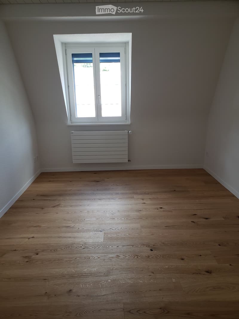 Empty room with wooden flooring, white walls, a window with white frame, and a white radiator below the window.