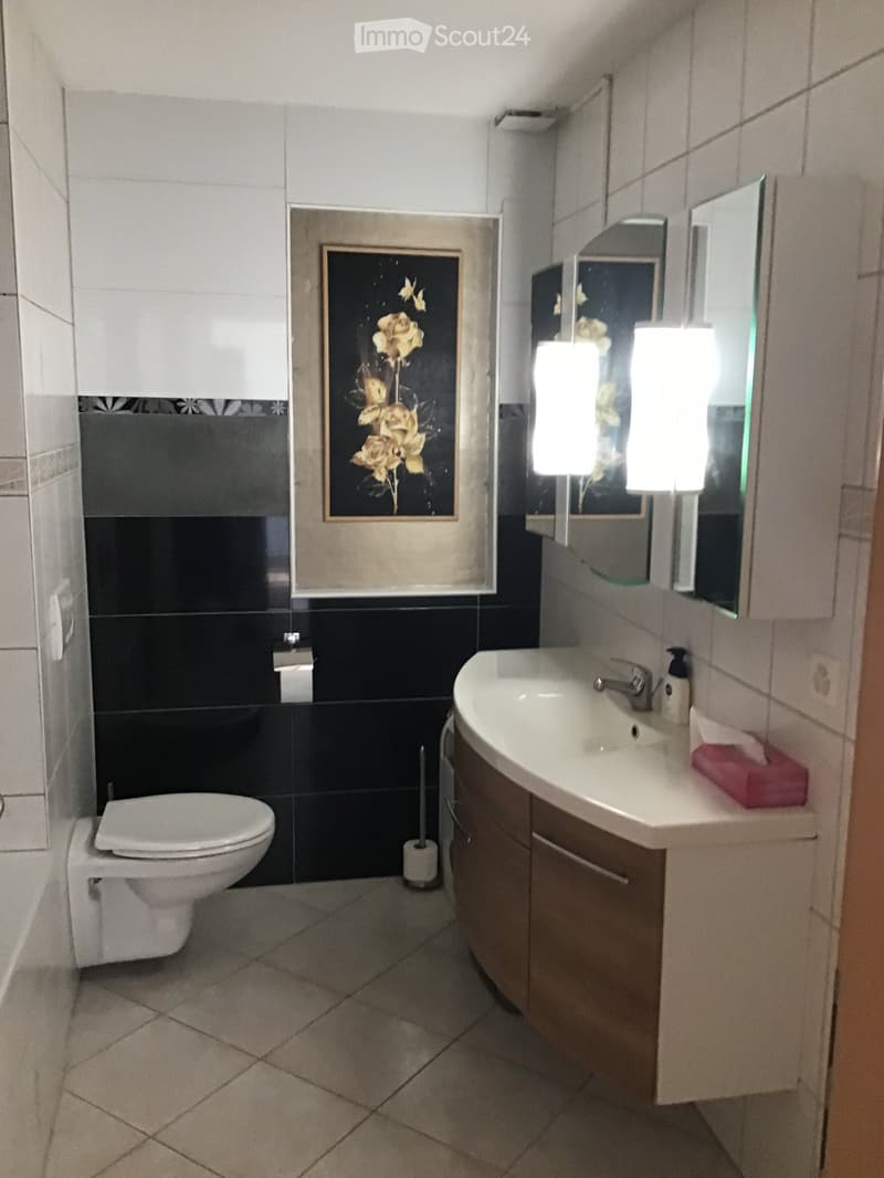 Modern bathroom with white tiles, black accents, a toilet, a sink with a mirror, and a picture frame on the wall.