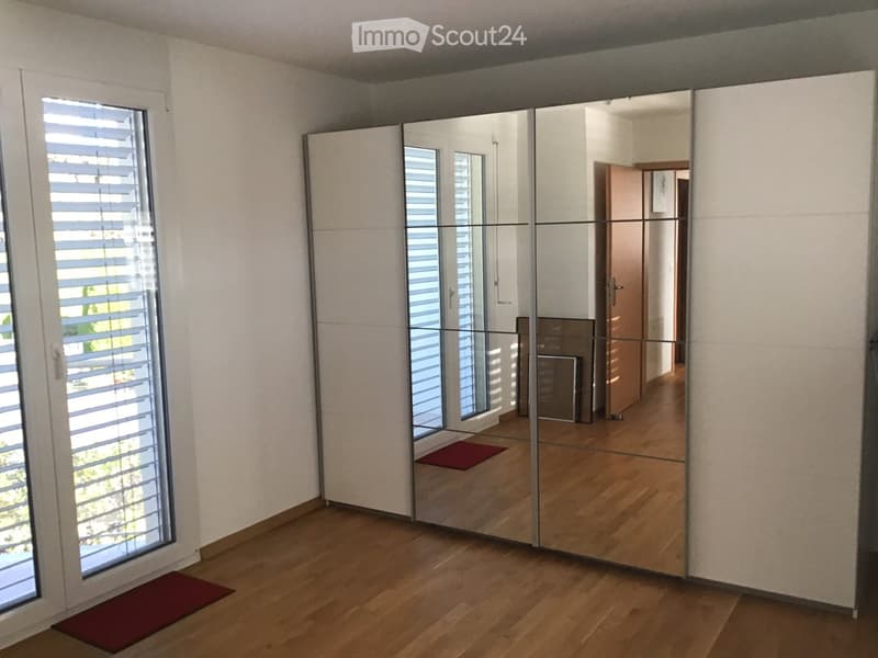 Empty room with wooden flooring, large mirrored wardrobe, and a window with blinds. The space is clean and unfurnished.