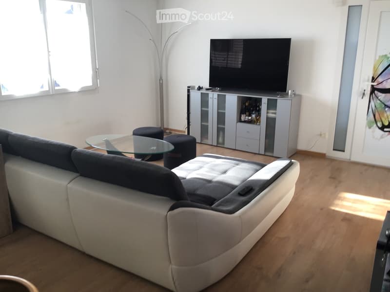Modern living room with a grey sectional sofa, glass coffee table, two black stools, TV stand with glass doors, and a floor lamp near the window.