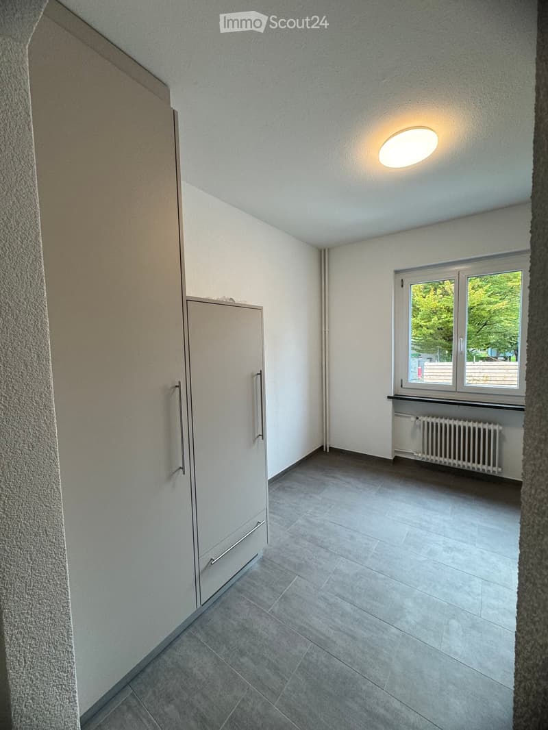 Empty room with grey tiled floor, white walls, built-in closet with mirrored doors, window with view of greenery, radiator, and ceiling light fixture.