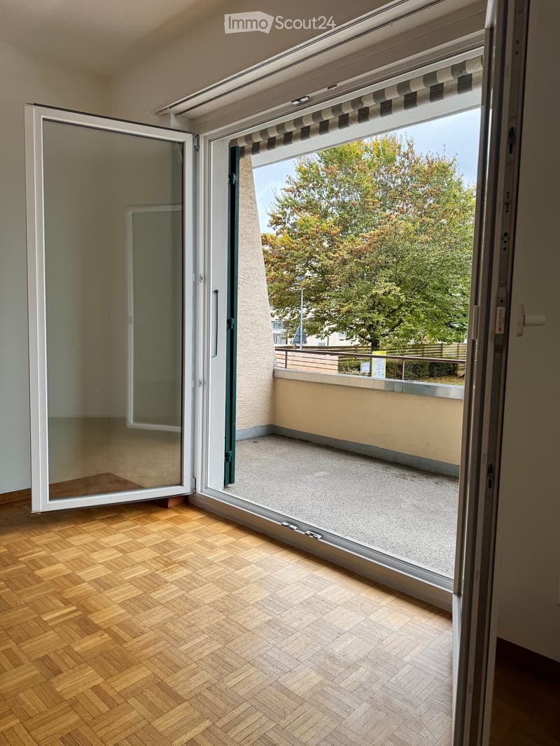 Open sliding glass doors lead to a small balcony with a concrete floor and a view of trees and a fenced area.