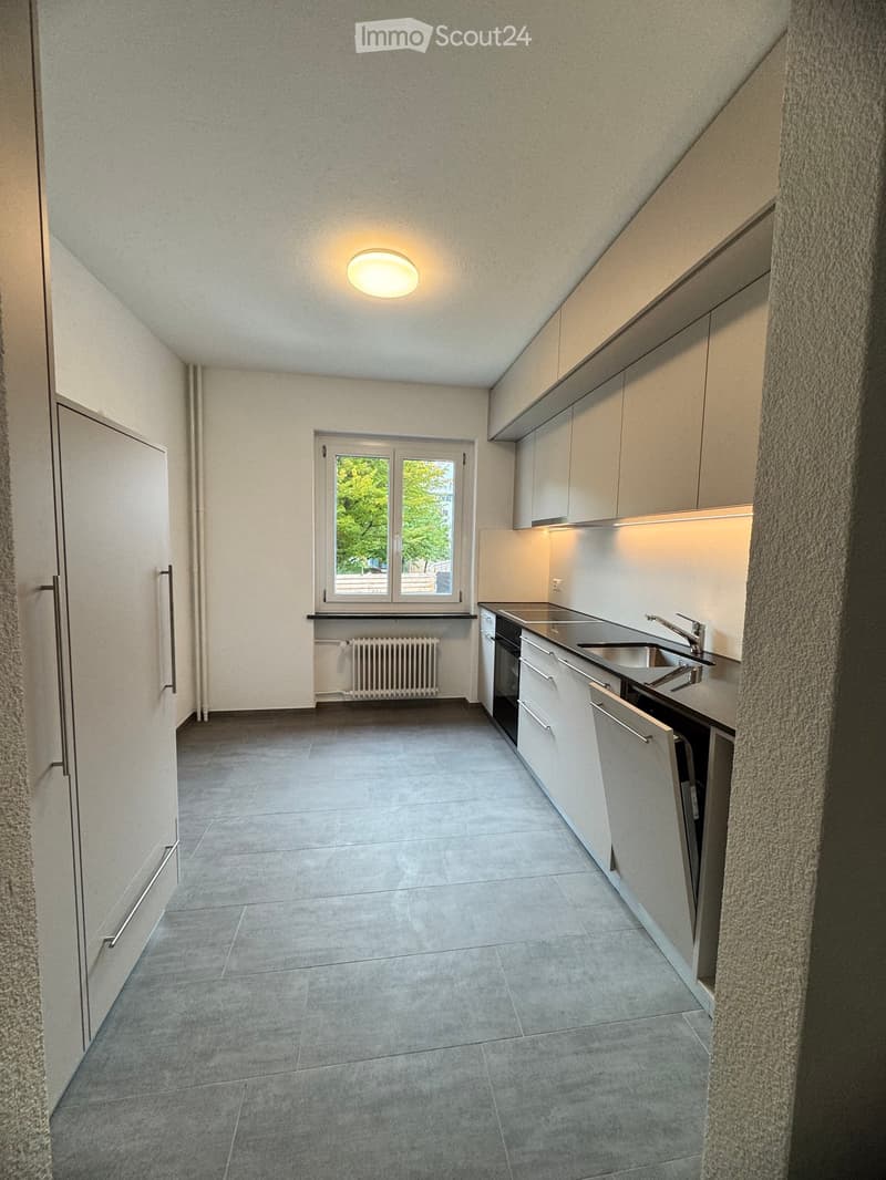 Modern kitchen with white cabinets, stainless steel sink, and built-in oven. Features a large window with a view of greenery, tiled floor, and a radiator under the window.