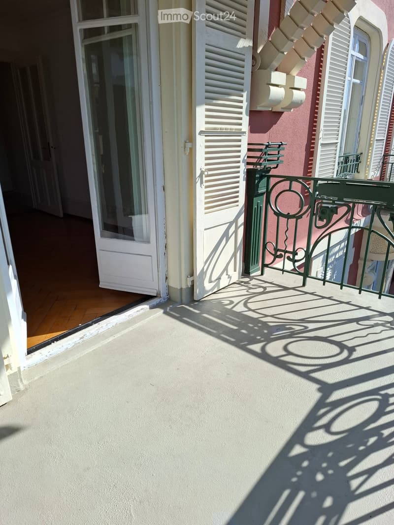Balcony with white walls, green metal railing, and shadows cast by nearby structures.