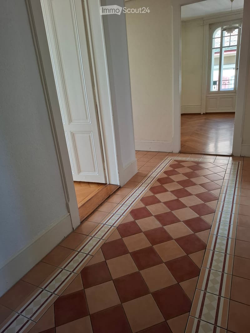 A hallway with patterned tiles, white walls, and a wooden floor visible in the adjacent room. The hallway features a white door on the left and a large window at the end, allowing natural light to enter.